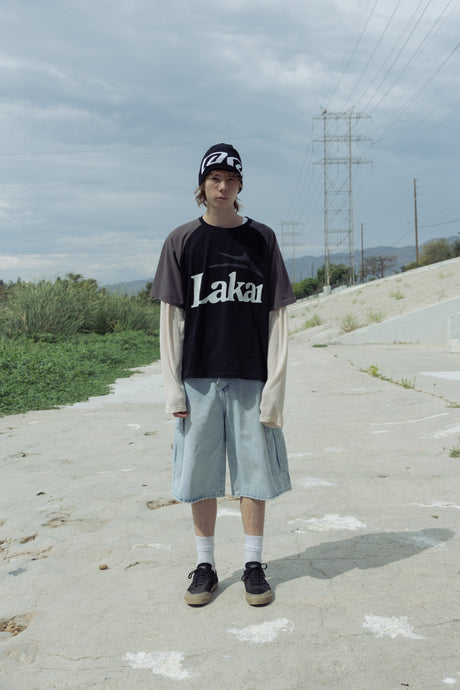 Person wearing a black 'Laka' t-shirt and light blue shorts standing on a road with power lines in the background.